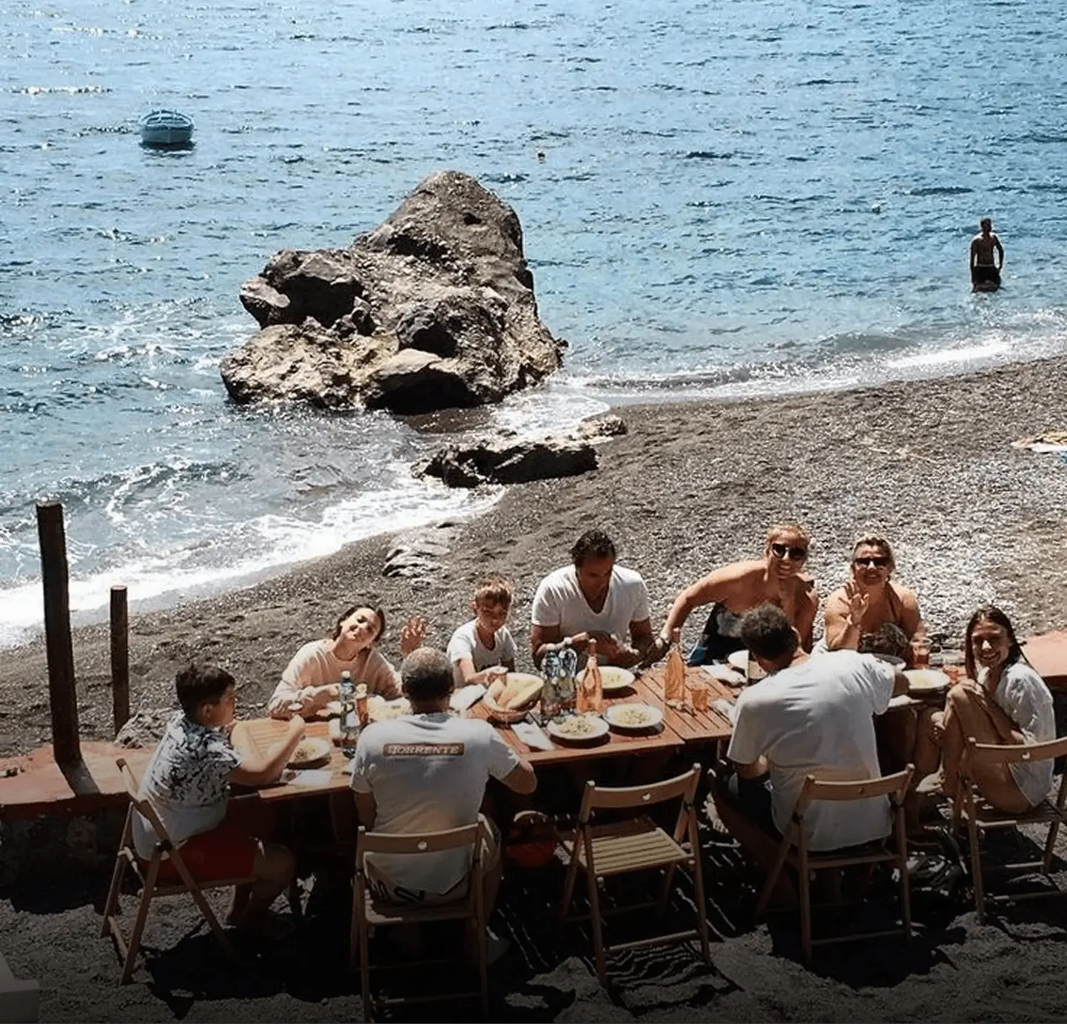 Grupo de personas compartiendo una comida en una mesa de madera ubicada en una playa de arena oscura, con el mar y una gran roca de fondo en un día soleado.